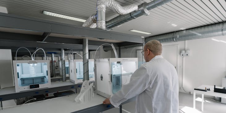 A man in white lab coat watching a 3D printer at work.