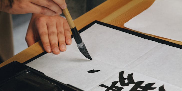 A hand holding a brush painting calligraphy with black ink on white paper.