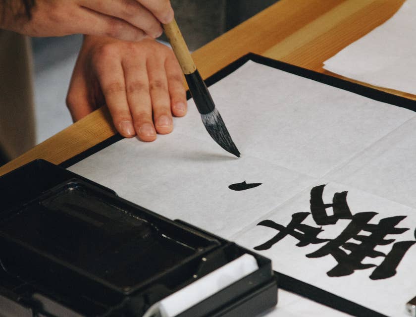 A hand holding a brush painting calligraphy with black ink on white paper.