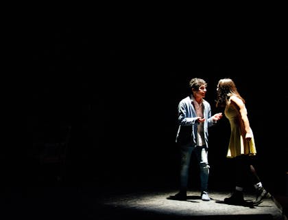 Dois atores no palco realizando uma cena dramática sob os holofotes em uma escola de teatro.