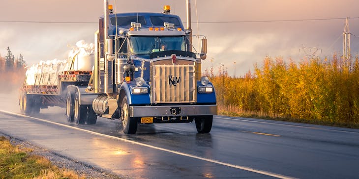 Blue truck with an open trailer transporting white bags secured with straps on a rainy rural road.