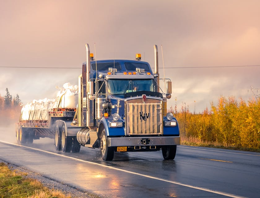 Camion bleu avec une remorque ouverte transportant des sacs blancs fixés par des sangles sur une route de campagne pluvieuse.