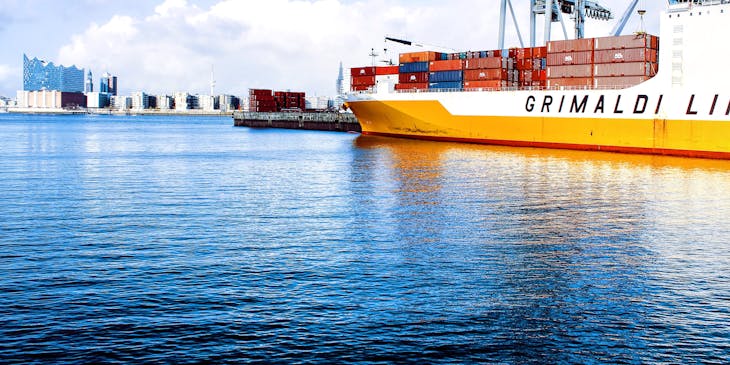 Harbor scene with cargo ship loaded with stacked containers docked at a harbor with a container crane in the background.