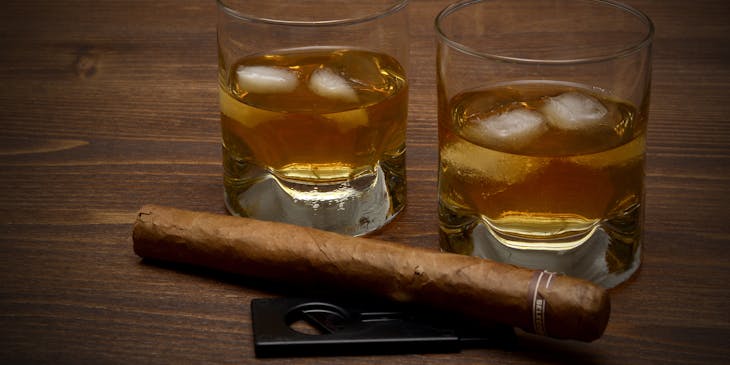 Two glasses of whiskey with ice, a cigar, and a cigar cutter set on a table in a cigar bar.