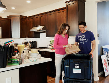 Eine Frau mit weißer Hose und pinkem Oberteil steht in ihrer Küche neben einem Mitarbeiter einer Entrümpelungsfirma, umgeben von Boxen und ausgeräumten Haushaltswaren.