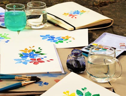 Varias pinturas de acuarela y pinceles entre un tintero y vasos servidos con agua en un negocio de pintura con vino.
