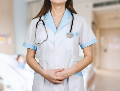 Eine Mitarbeiterin eines Krankenhauses mit einem Stethoskop trägt ihre weiße Arbeitskleidung, die mit hellblauen Rändern an den Armen und am Halsausschnitt abgesetzt ist.