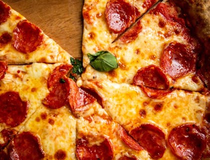 Una pizza a fette con salame sopra un tagliere di legno tipico delle pizzerie al taglio.