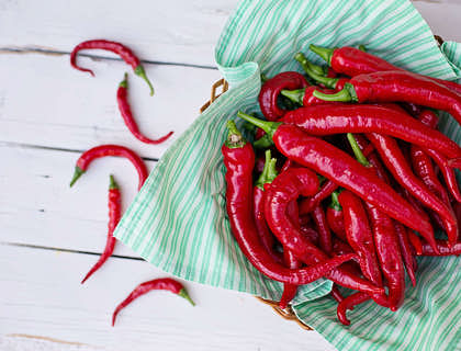Dei peperoncini rossi in un cesto sopra un panno verde acqua che si trova sopra un tavolo di legno bianco.