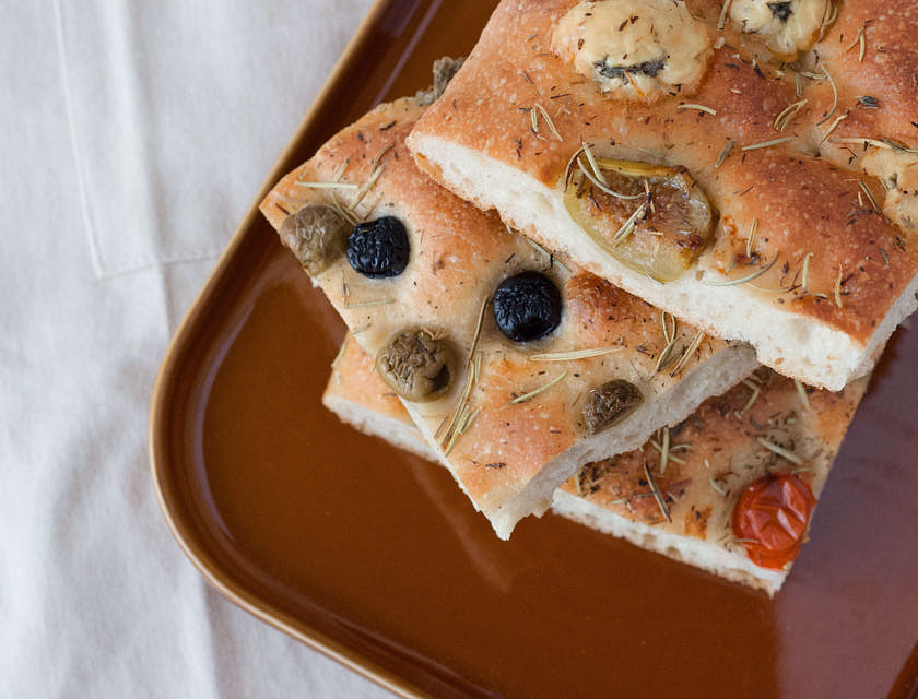 Tre fette di focaccia pugliese in un piatto marrone sopra un tavolo con una tovaglia bianca.