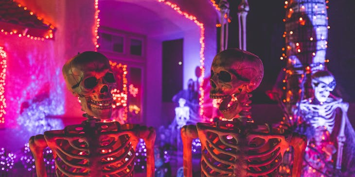 Two smiling skeletons looking at each other in front of a colorfully lit haunted mansion.