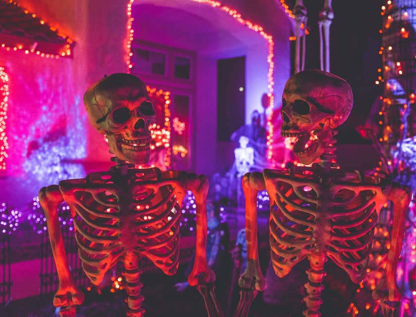 Two smiling skeletons looking at each other in front of a colorfully lit haunted mansion.