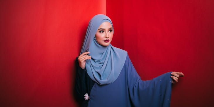 Woman wearing a blue hijab and dress against a red wall.