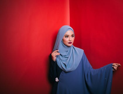 Una mujer vistiendo un hijab azul y vestido contra una pared roja en un negocio de hijabs.