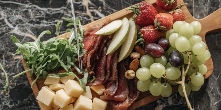 A wooden grazing board with cold cuts, cheese cubes, fresh herbs, and slices of fresh fruit.