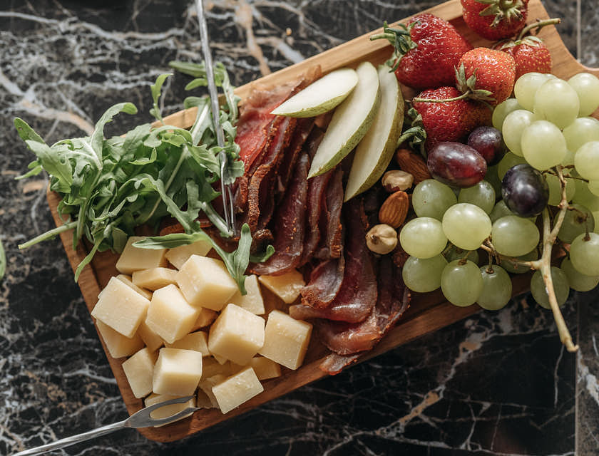 Une planche apéritive en bois avec de la charcuterie, des cubes de fromage, des herbes fraîches et des fruits frais.