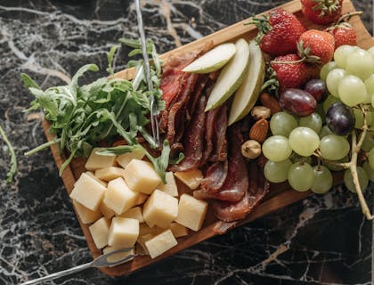 A wooden grazing board with cold cuts, cheese cubes, fresh herbs, and slices of fresh fruit.