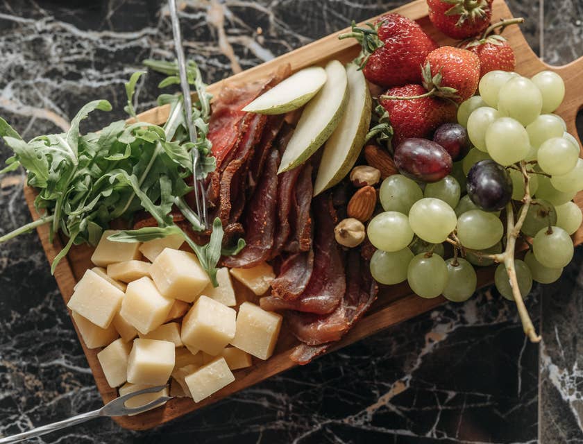 Uma tábua de madeira com frios, cubos de queijo, ervas e frutas frescas.