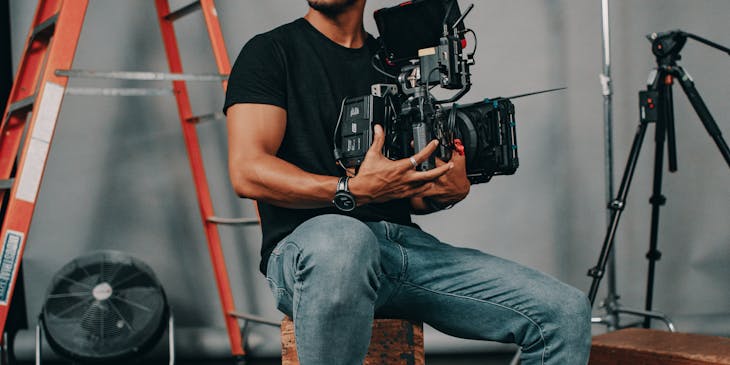 Miembro de un equipo de filmación sentado en una caja de madera sosteniendo una cámara de video de una empresa productora.