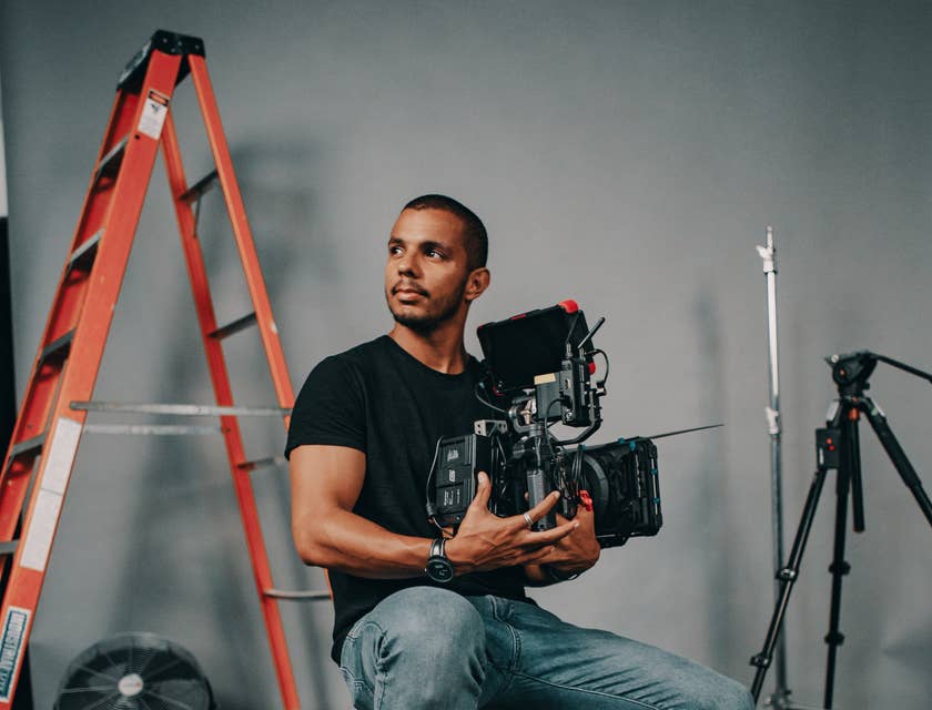 Miembro de un equipo de filmación sentado en una caja de madera sosteniendo una cámara de video de una empresa productora.