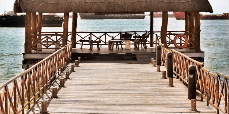 Palapa a la orilla del mar con mesas y sillas, un muelle de entrada y barcos al fondo.