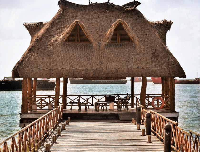 Palapa a la orilla del mar con mesas y sillas, un muelle de entrada y barcos al fondo.