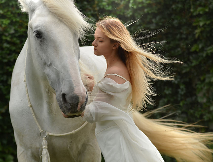 Una attraente donna bionda vestita di bianco che accarezza un cavallo bianco.