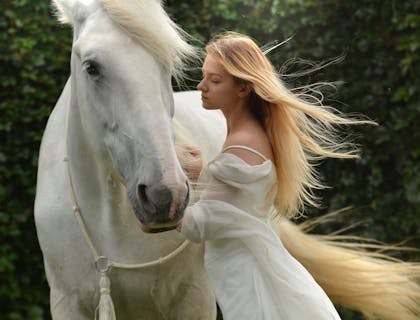 Una attraente donna bionda vestita di bianco che accarezza un cavallo bianco.