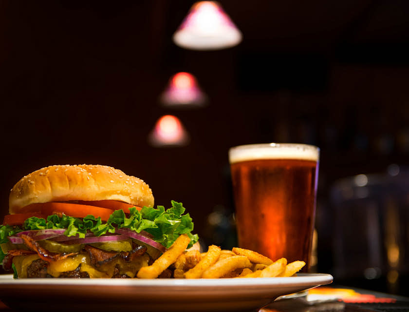Una hamburguesa con papas a la francesa en un plato a un lado de una cerveza oscura en una hamburguesería.