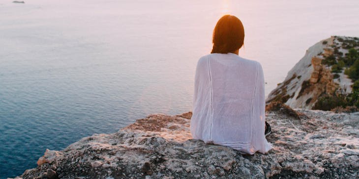 A woman sitting on a rock overlooking a calm ocean scene.