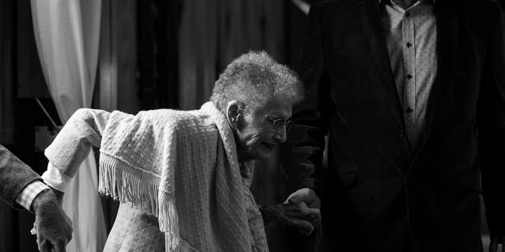 Two smartly-dressed caregivers helping an elderly woman onto her feet.