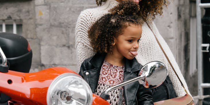 A mother and daughter sticking tongues out in the mirror of a motorcycle.