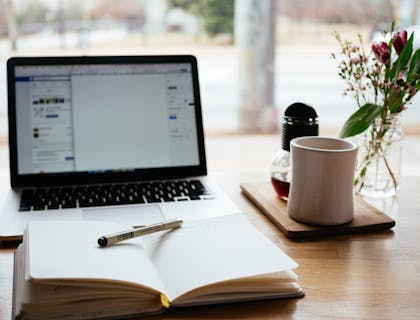una libreta abierta con un lápiz en medio frente a una laptop y a un lado de una taza blanca con café en un negocio creativo