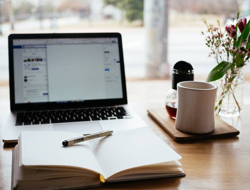 una libreta abierta con un lápiz en medio frente a una laptop y a un lado de una taza blanca con café en un negocio creativo