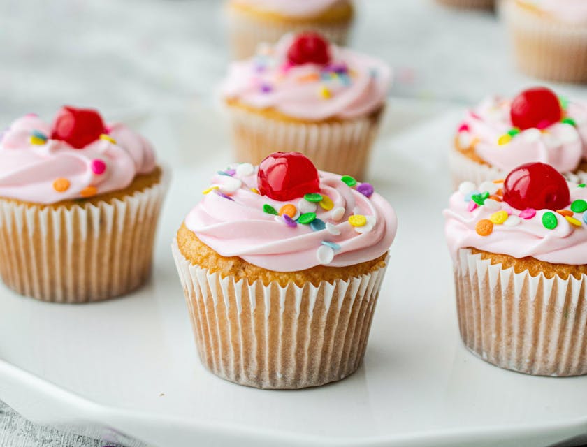 Cupcakes con betún rosado con cerezas sobre un plato de cerámica blanca en una superficie gris en un negocio de cupcakes.