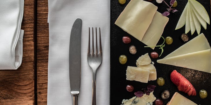 Cutlery, including a knife and a fork, placed on a white linen napkin on a table between a plate with an assortment of cheeses.