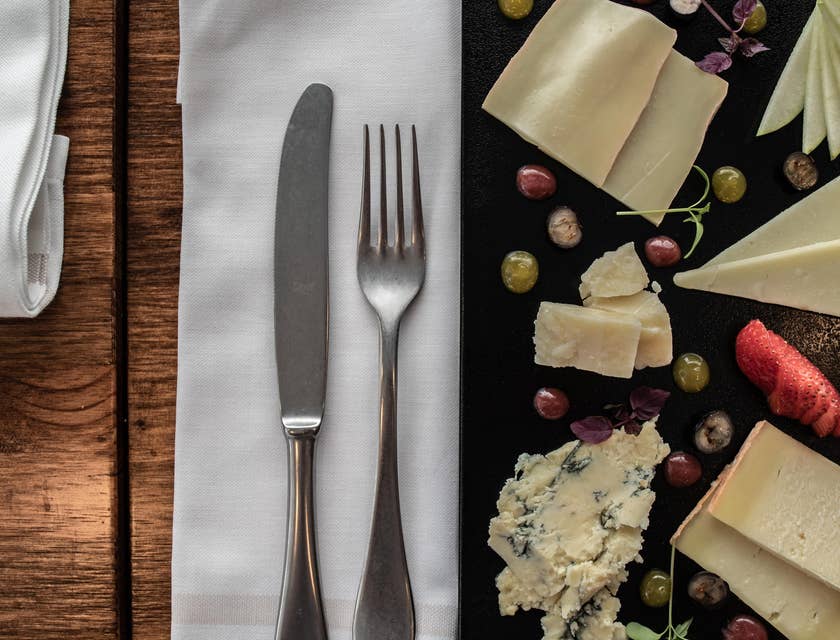 Cutlery, including a knife and a fork, placed on a white linen napkin on a table between a plate with an assortment of cheeses.