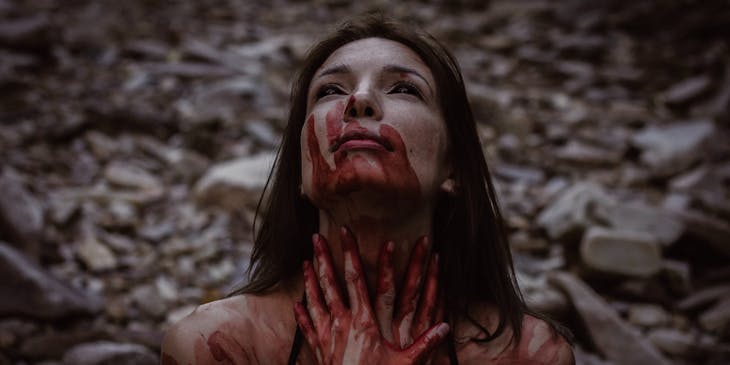 woman with black eyes and blood-smeared face, neck, and hands in a dark scene