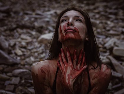 woman with black eyes and blood-smeared face, neck, and hands in a dark scene