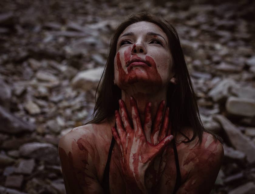 woman with black eyes and blood-smeared face, neck, and hands in a dark scene