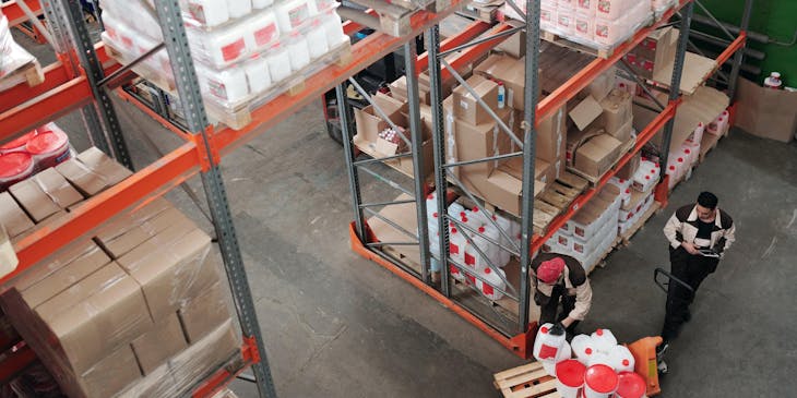 Two people in a depot transporting goods on a wooden trolley.
