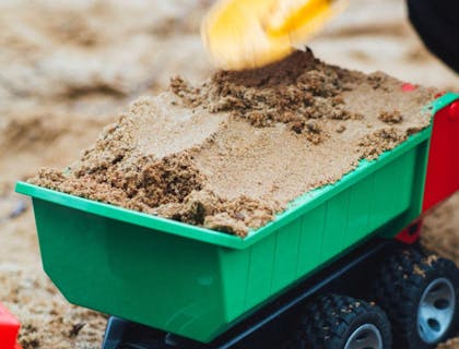 Close-up of a child's hand holding a spade and filling a toy dump trailer with a load of sand.