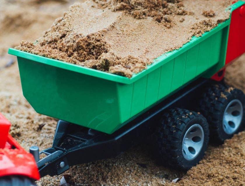 Close-up of a child's hand holding a spade and filling a toy dump trailer with a load of sand.