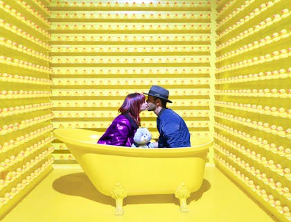 Dos personas besándose en una bañera amarilla, rodeada de patos de goma amarillos en un negocio con nombre vanguardista.