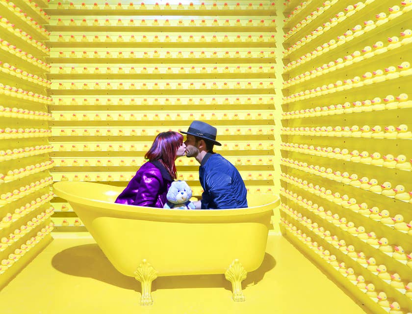 Dos personas besándose en una bañera amarilla, rodeada de patos de goma amarillos en un negocio con nombre vanguardista.