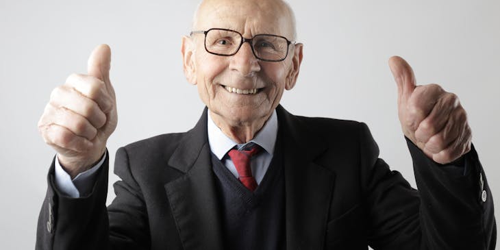 Elderly man in a black suit giving thumbs up signs with both hands.