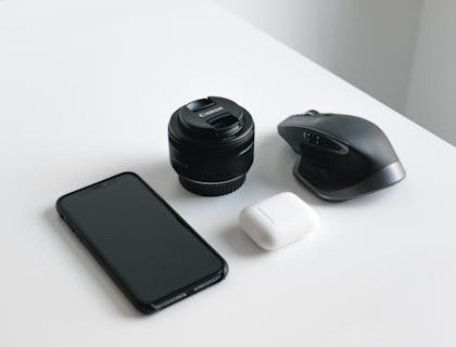 Teléfono móvil, lente de cámara digital y mouse inalámbrico acomodados sobre una mesa blanca en un negocio de electrónicos.