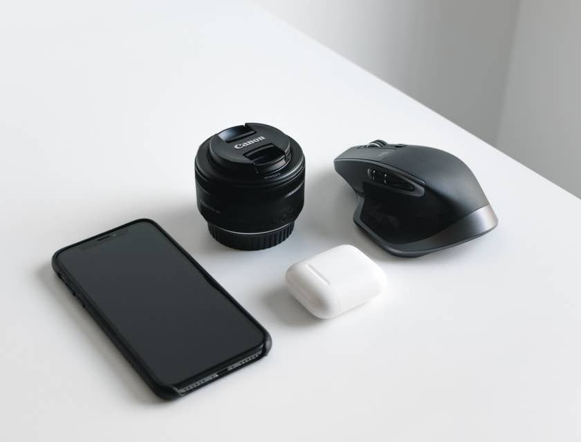 Smartphone preto, lente de câmera e mouse sem fio em cima de uma mesa branca.
