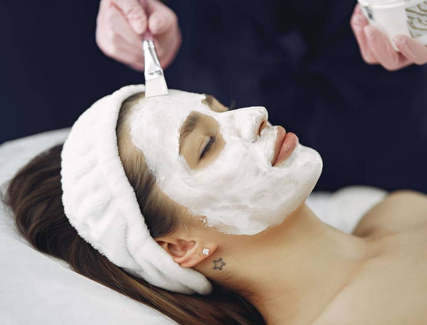 Eine Frau entspannt auf einer Liege mit geschlossenen Augen in einem Kosmetikstudio während eine weiße Creme mit einem Pinsel auf ihrem Gesicht aufgetragen wird.