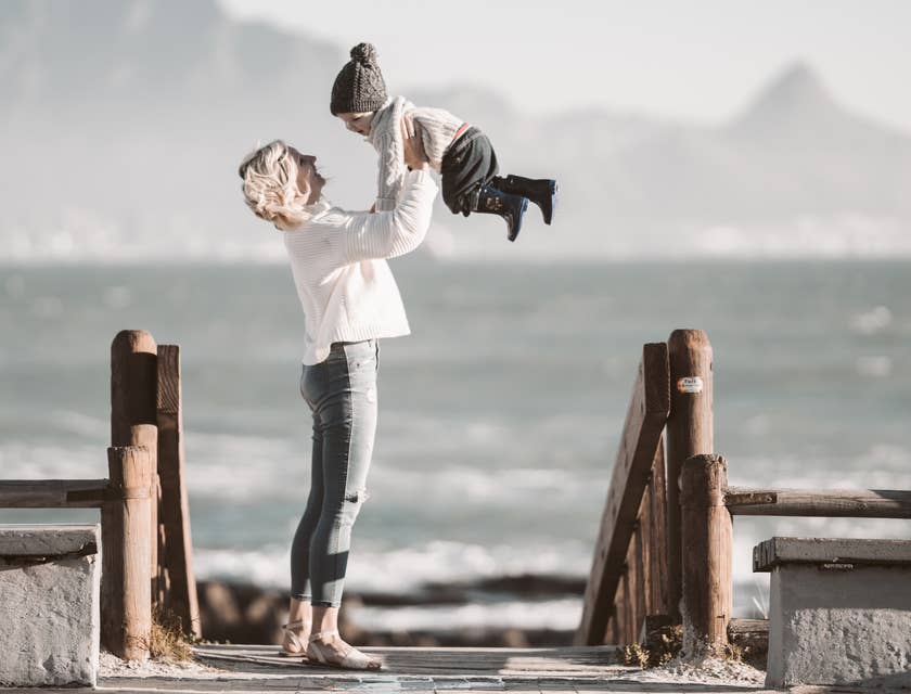una mamá cargando a su hijo en frente del mar en una playa durante una sesión de fotos de fotografía familiar